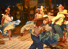 A fotografia mostra um grupo de pessoas dançando carimbó. O primeiro plano mostra um rapaz usando chapéu de palha, calça azul e blusa branca com babados, fazendo uma passada da dança, da esquerda para a direita. Ao fundo, mulheres vestindo saias azuis floridas, blusas brancas de babados e tiaras de flores rosa nos cabelos dançam com rapazes vestidos da mesma forma que o primeiro. No fundo da imagem, é possível ver o público que assiste à apresentação.